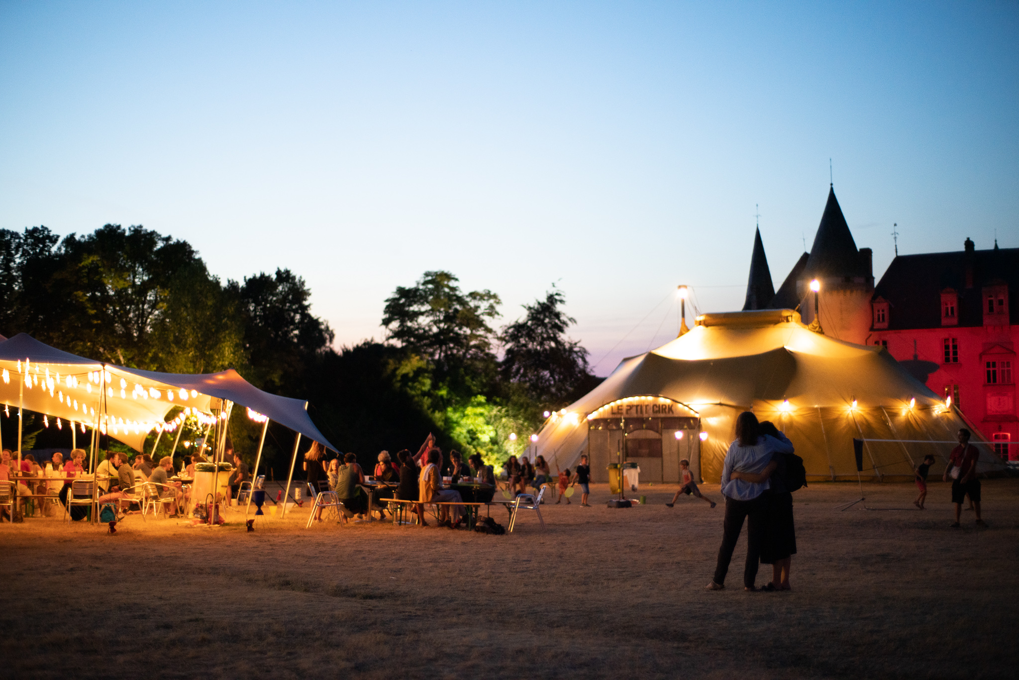 A la tombée de la nuit, au premier plan à droite, un chapiteau gris entouré d’une guirlande de lumières situé devant un château dont on aperçoit la façade et la toiture de deux tours. A gauche, une toile blanche tendue éclairée, derrière elle, un bosquet d’arbres. Devant, sur la pelouse, plusieurs personnes sont debout.