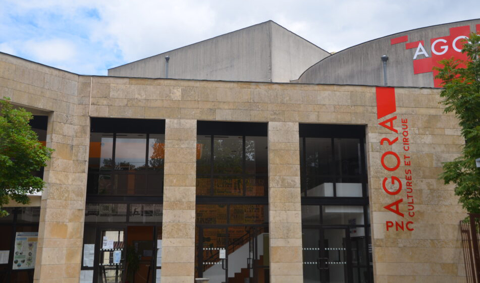 Façade d’un bâtiment massif en pierre claire percée de quatre grandes baies vitrées verticales. A droite est décrit verticalement en rouge sur deux lignes « Agora Cultures et cirque » et horizontalement « PNC ». Au-dessus on aperçoit le haut de l’enceinte de la salle de spectacle sur lequel est apposé le logo rouge de l’Agora.