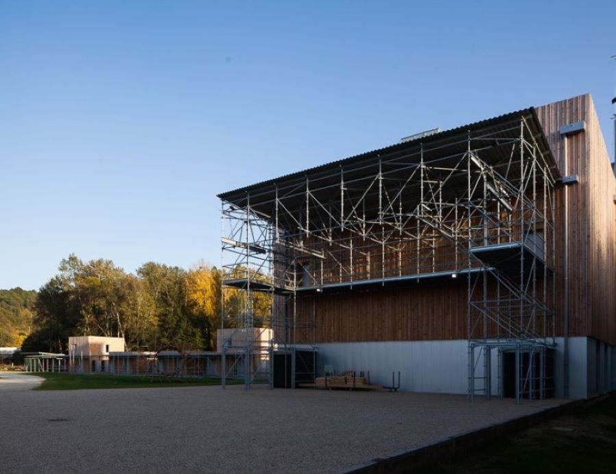 Devanture d'un bâtiment de forme cubique dont la façade est composée d’un bardage en bois et d’une bande horizontale en inox. Agrémentée d’un échafaudage décoratif en acier galvanisé. Le bâtiment est implanté sur des graviers et entouré de pelouse. Deux portes d’entrées marrons se trouvent de chaque côté (droit et gauche). Sur la gauche du bâtiment, une lignée d’appartements en bois avec échafaudages.