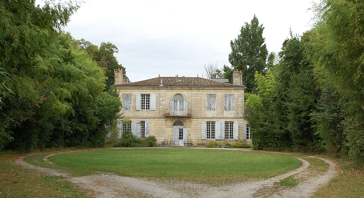 La photo montre une grande maison de pierre claire à deux étages, de forme rectangulaire, avec un toit bas en tuiles brunes et quatre cheminées. La façade est symétrique, percée de hautes fenêtres à volets gris, certaines fermées. Au centre, une porte surmontée d’une grande fenêtre cintrée et d’un petit balcon en fer. La maison est entourée d’arbres denses et un chemin de gravier forme un cercle devant une pelouse verte.