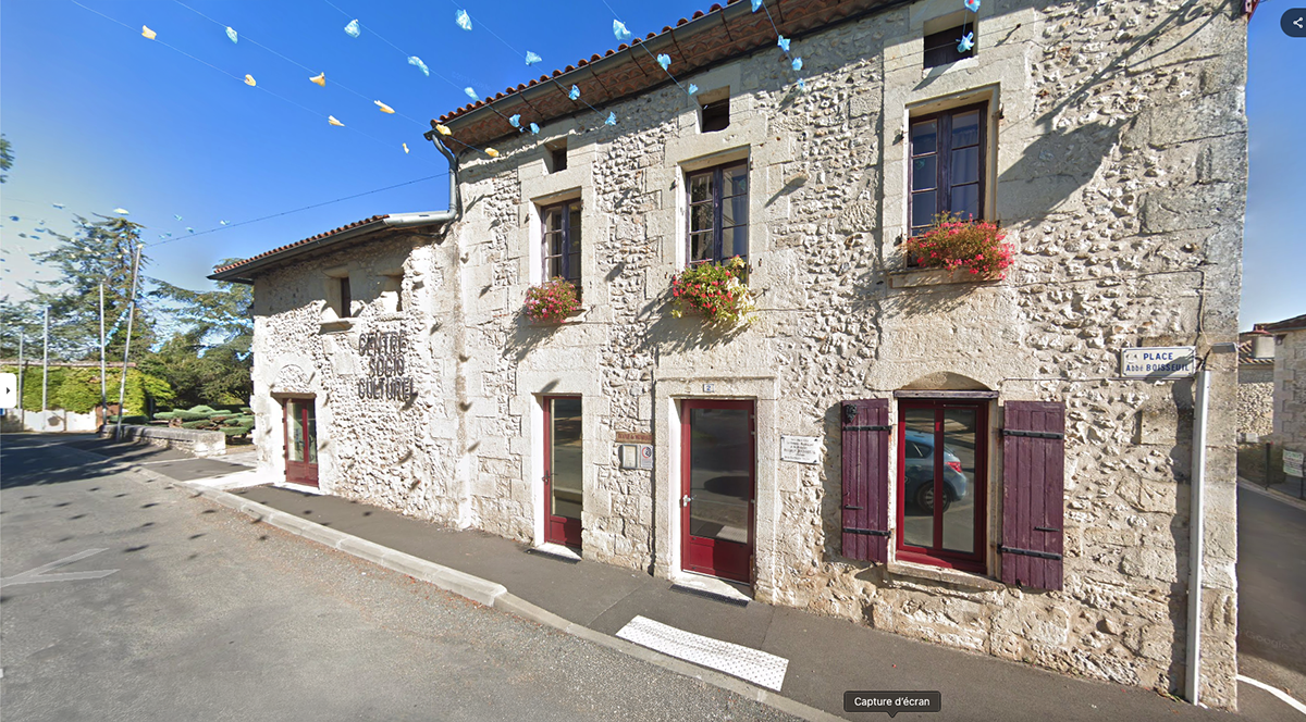 Le bâtiment est en pierre claire, avec des murs irréguliers typiques d’une construction ancienne. Il est sur deux niveaux, couvert d’un toit de tuiles rouges. En façade, plusieurs portes vitrées encadrées de bois sombre mènent directement sur la rue, certaines avec volets en bois peints en bordeaux. À l’étage, des fenêtres rectangulaires sont ornées de jardinières fleuries. Une enseigne en relief sur la pierre indique “Centre Socio Culturel”. L’ensemble donne une impression solide et rustique, occupant l’angle d’une petite place.