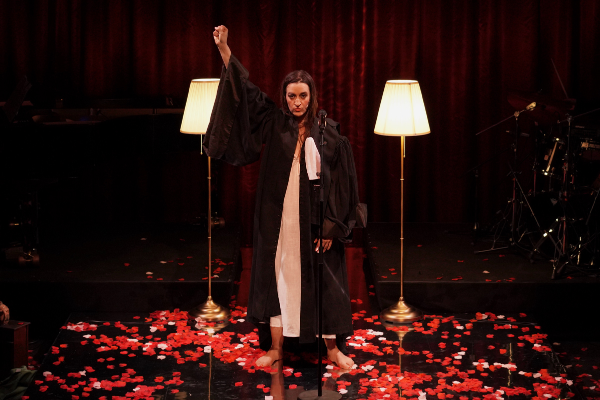 Au centre d’une scène au sol jonché de confettis rouges et blancs, une femme debout en tenue de juriste, longue robe noire ouverte sur une longue robe blanche, lève le poing. Elle regarde le public. Derrière elle, de part et d’autre, deux lampadaires aux pieds dorés et abat-jour blancs sont allumés. Derrière elle, en fond de scène, un rideau rouge apparaît.